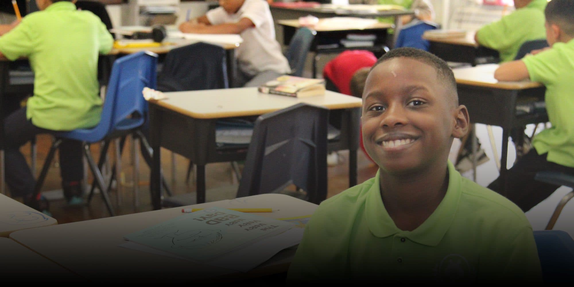 Smiling student in a green uniform shirt at a classroom desk.